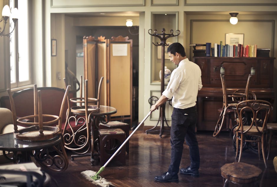 A professional cleaner dressed in a white protective suit, face mask, and blue gloves is performing surface cleaning on a large white cushion or mattress with a handheld vacuum or steamer. The room fe